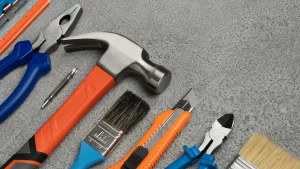 Ferramentas manuais como martelo, alicates, estilete, pincéis e bits dispostas sobre superfície cinza, representando a importância de escolher a ferramenta certa para cada tipo de trabalho.